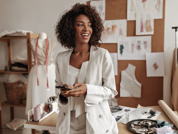 Happy charming dark-skinned curly brunette woman in white jacket, stylish top smiles sincerely, looks away, holds phone. Fashion designer poses in office.